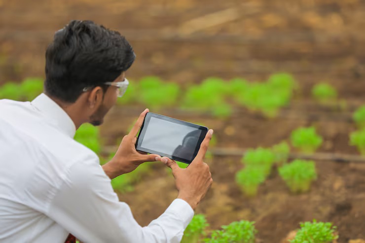 A young Indian agronomist in a white shirt uses a tablet to monitor crop health in a field with green plants. This represents smart farming using VYOM GIS technology.